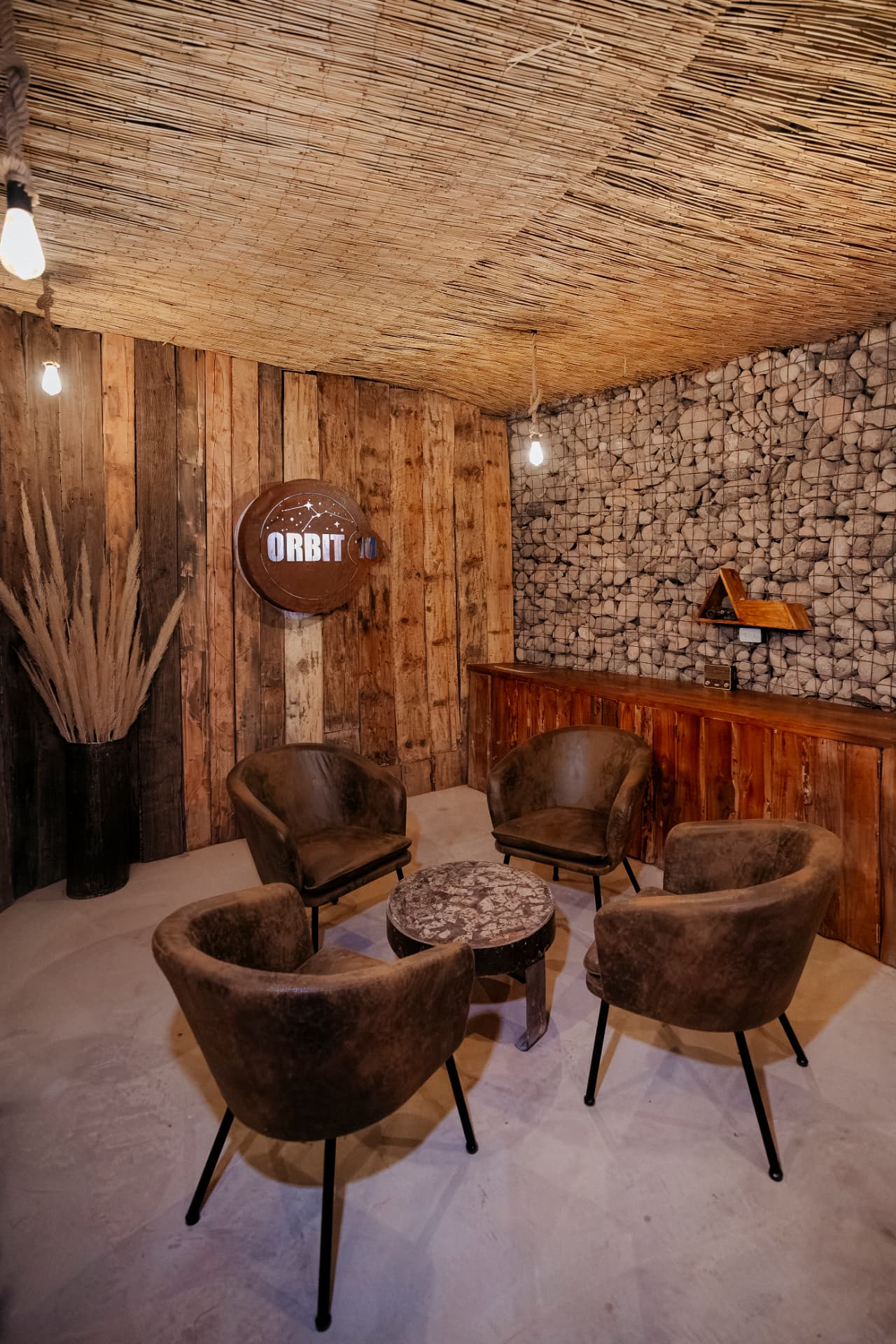 Lounge interno do Tour Astronômico - Orbit 10 com poltronas, parede de pedras e teto de fibra natural