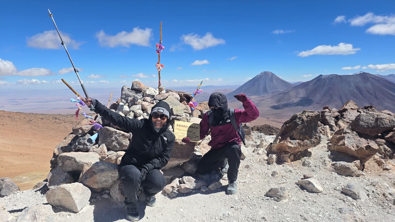 Dois viajantes comemoram no cume do Cerro Toco, com placas de altitude e vulcões andinos ao fundo sob céu azul