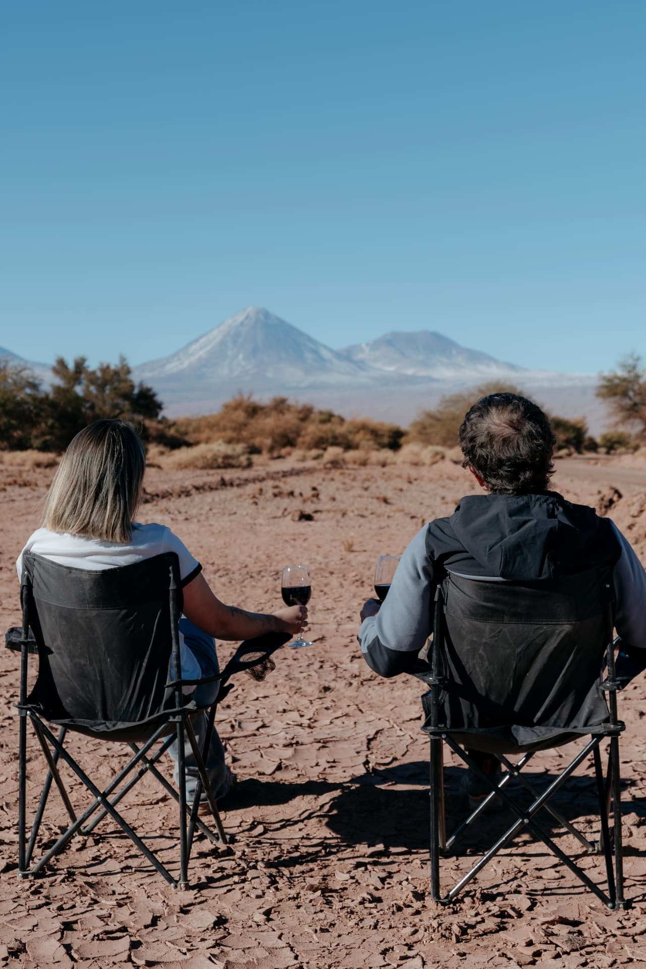 Casal sentado em cadeiras no deserto brindando com vinho diante do vulcão Licancabur