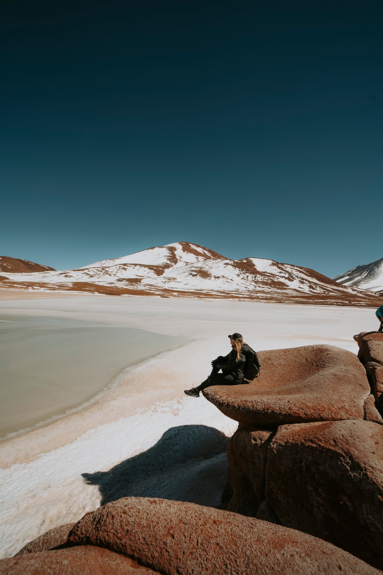 Viajante sentada em rocha diante de laguna altiplânica nevada