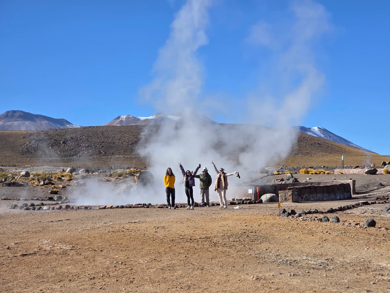 Grupo de viajantes comemora diante das colunas de vapor dos Gêiseres del Tatio, com vulcões nevados ao fundo