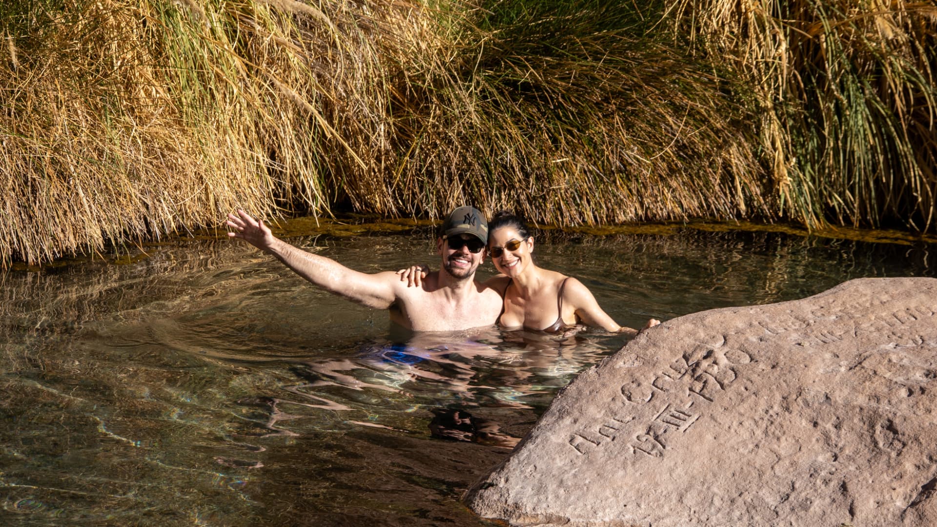 Casal relaxando nas águas cristalinas das Termas de Puritama, cercado por vegetação do deserto no Atacama